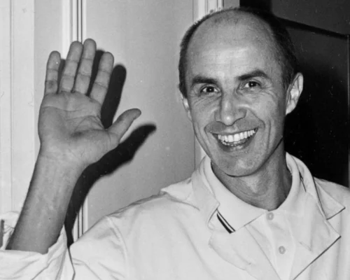 Fotografía en blanco y negro de un hombre sonriente, con bata blanca, levantando la mano en gesto de saludo frente a una puerta en un interior.