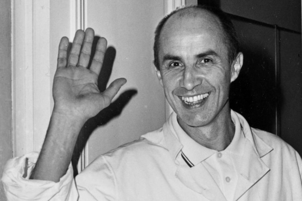 Fotografía en blanco y negro de un hombre sonriente, con bata blanca, levantando la mano en gesto de saludo frente a una puerta en un interior.